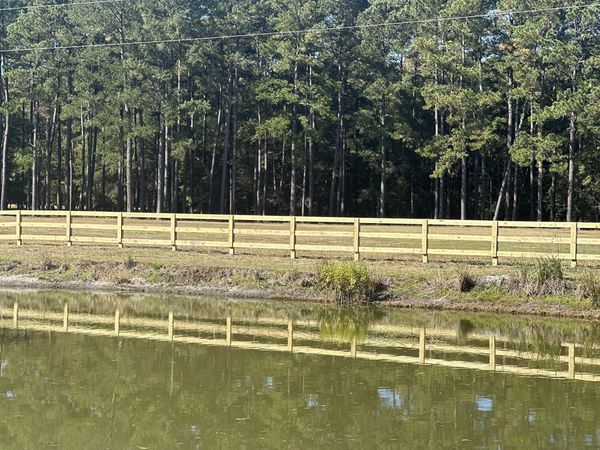 A picturesque 3-rail wood fence bordering a serene pond with a backdrop of lush pine trees.